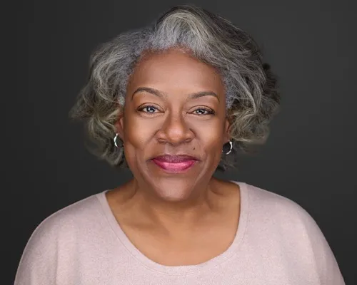 Warm, confident headshot of a woman with silver-gray hair, magenta lipstick, and taupe crew neck shirt against dark gray studio backdrop