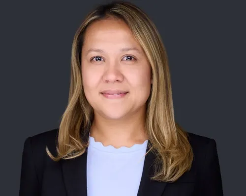 Professional headshot of woman in black blazer and light blue shirt with blonde highlights against neutral dark blue-gray studio backdrop