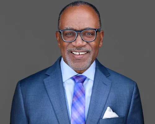 Professional headshot of man with clear-framed glasses, gray-tempered hair, blue blazer and striped tie against neutral gray studio backdrop