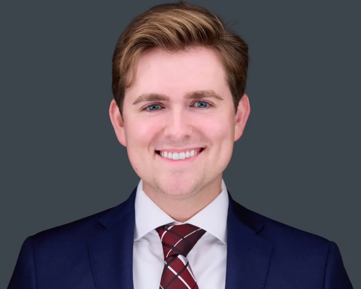 Male corporate headshot with confident smile wearing navy blazer, white shirt, and burgundy plaid tie