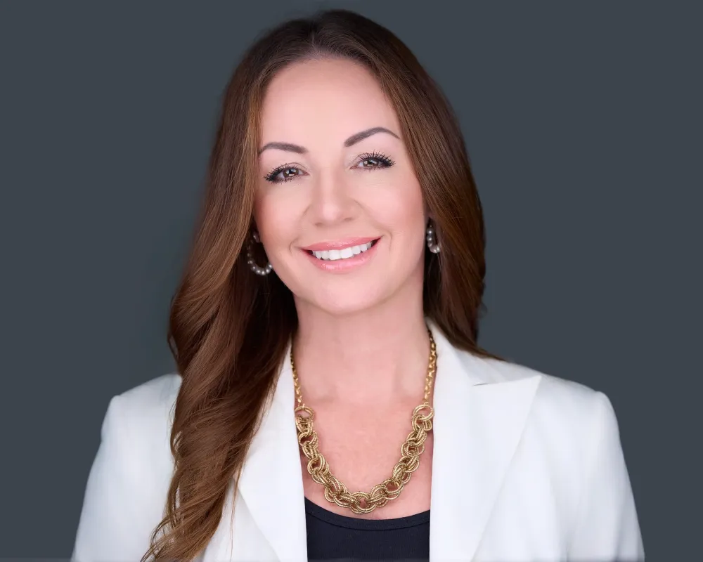 Jennifer Dennis professional headshot in white blazer with gold jewelry displaying confident warm smile against gray studio backdrop