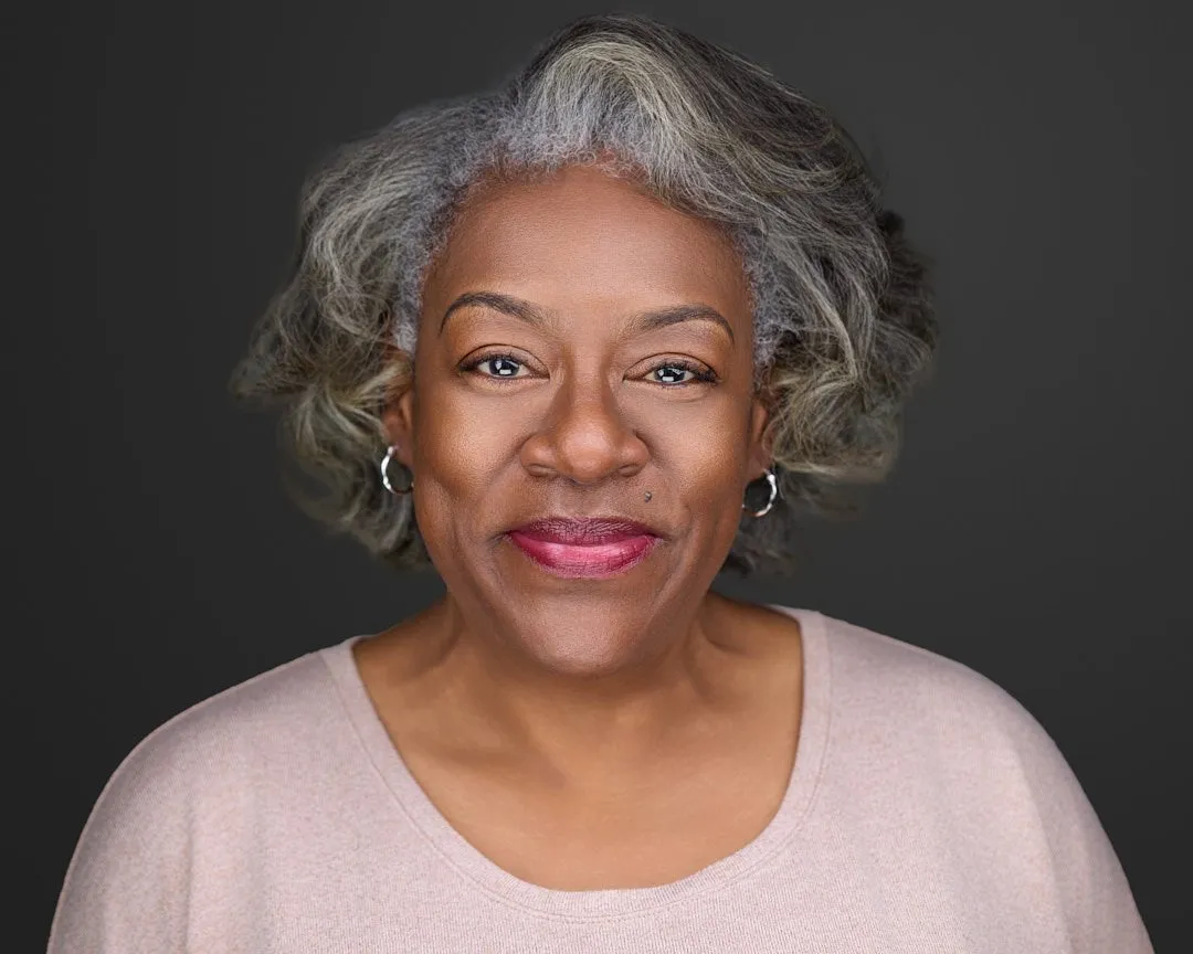 Professional headshot of mature woman with gray curly hair and bold magenta lipstick against dark background