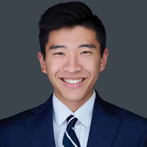Professional male realtor headshot in navy suit and striped tie against neutral gray studio background
