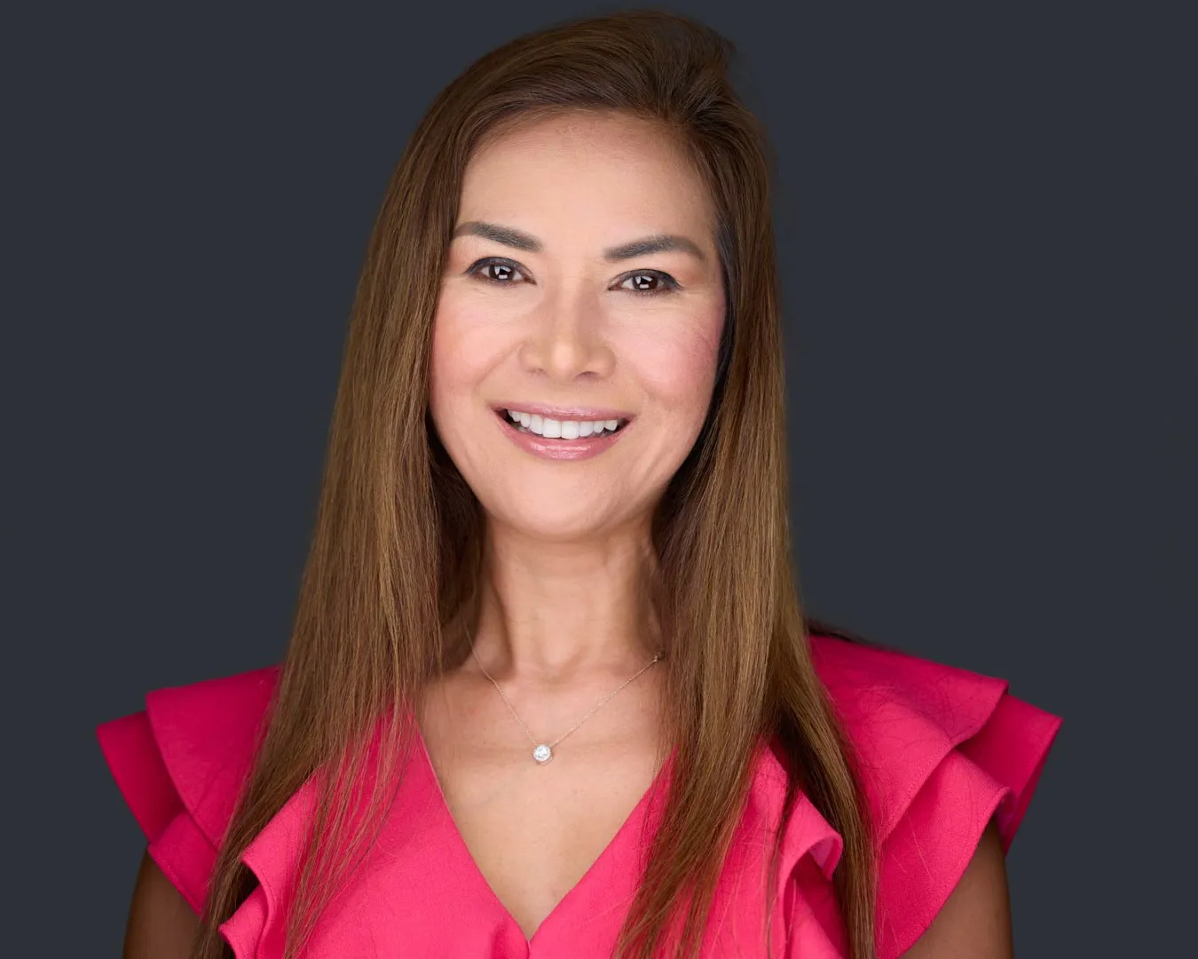 Gift Thongpia-Hughes professional headshot in vibrant pink top displaying genuine smile against neutral dark studio background